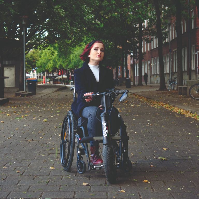 Luisa sitzt in ihrem Rollstuhl an dem ein Zuggerät montiert ist. Sie steht in einer leeren Allee und der Wind weht ihr durch die Haare.